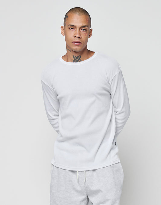 A person with a shaved head and visible neck and face tattoos wears the LIVE FAST DIE YOUNG Basic Regular Rib Longsleeve in white, paired with light-colored pants, standing against a plain white background with hands behind their back.