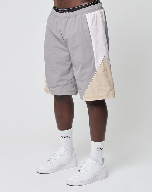 A person wearing LIVE FAST DIE YOUNG Basic Nylon Trackshorts in gray, white, and beige colorblock with a Regular Fit and elastisches Bündchen, plus white crew socks with LFDY text and white Nike Air Force 1s, stands against a plain white background.