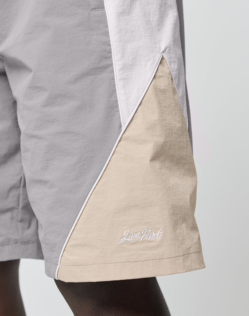 Close-up of a person wearing LIVE FAST DIE YOUNG Basic Nylon Trackshorts in light grey, beige, and white, featuring a Nylon-Lycra blend, elastic waistband, piping detail, and “Live Fast” embroidery near the hem.