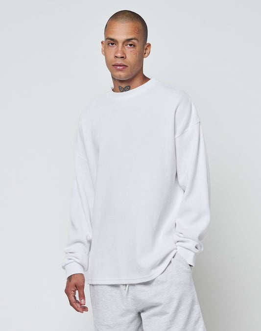 A person with a shaved head, face and neck tattoos wears the LIVE FAST DIE YOUNG Blank Waffle Longsleeve in white (oversize fit, 100% Baumwolle, Ripp-Bündchen) and light pants against a plain light gray background.