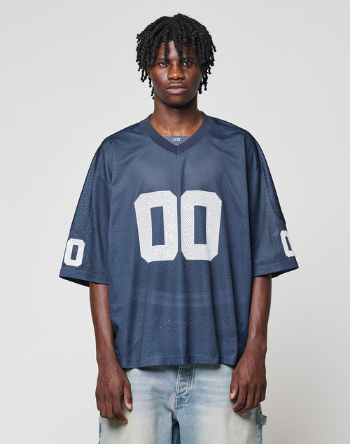 A man with short dreadlocks wears the LFDY Mesh Jersey by LIVE FAST DIE YOUNG in navy blue polyester, featuring a V-neck and white number 00 on front and sleeves, paired with light blue jeans against a plain light background.