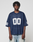 A person wears a loose-fitting LIVE FAST DIE YOUNG LFDY Mesh Jersey in navy blue with a V-neck and white number 00 on the front and sleeves, paired with light blue jeans, standing against a plain light gray background.