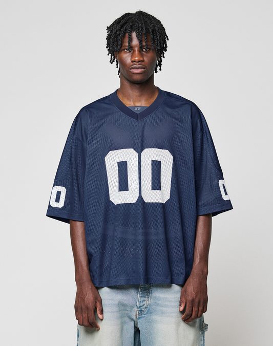 A person wears a loose-fitting LIVE FAST DIE YOUNG LFDY Mesh Jersey in navy blue with a V-neck and white number 00 on the front and sleeves, paired with light blue jeans, standing against a plain light gray background. | model