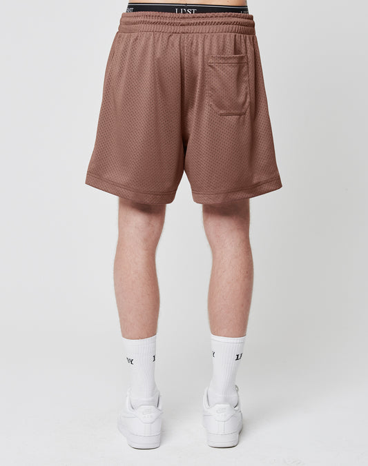 A person stands facing away, wearing LIVE FAST DIE YOUNG Basic Mesh Shorts in brown with a logo patch, elastic waistband, and back pocket, paired with white socks and sneakers. The background is plain and light-colored.