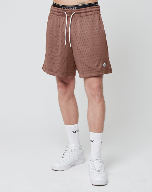 A person stands against a plain white background wearing LIVE FAST DIE YOUNG Basic Mesh Shorts in light brown, white socks with black text, white sneakers, and a black waistband with white lettering and a logo patch. The upper body is not visible.
