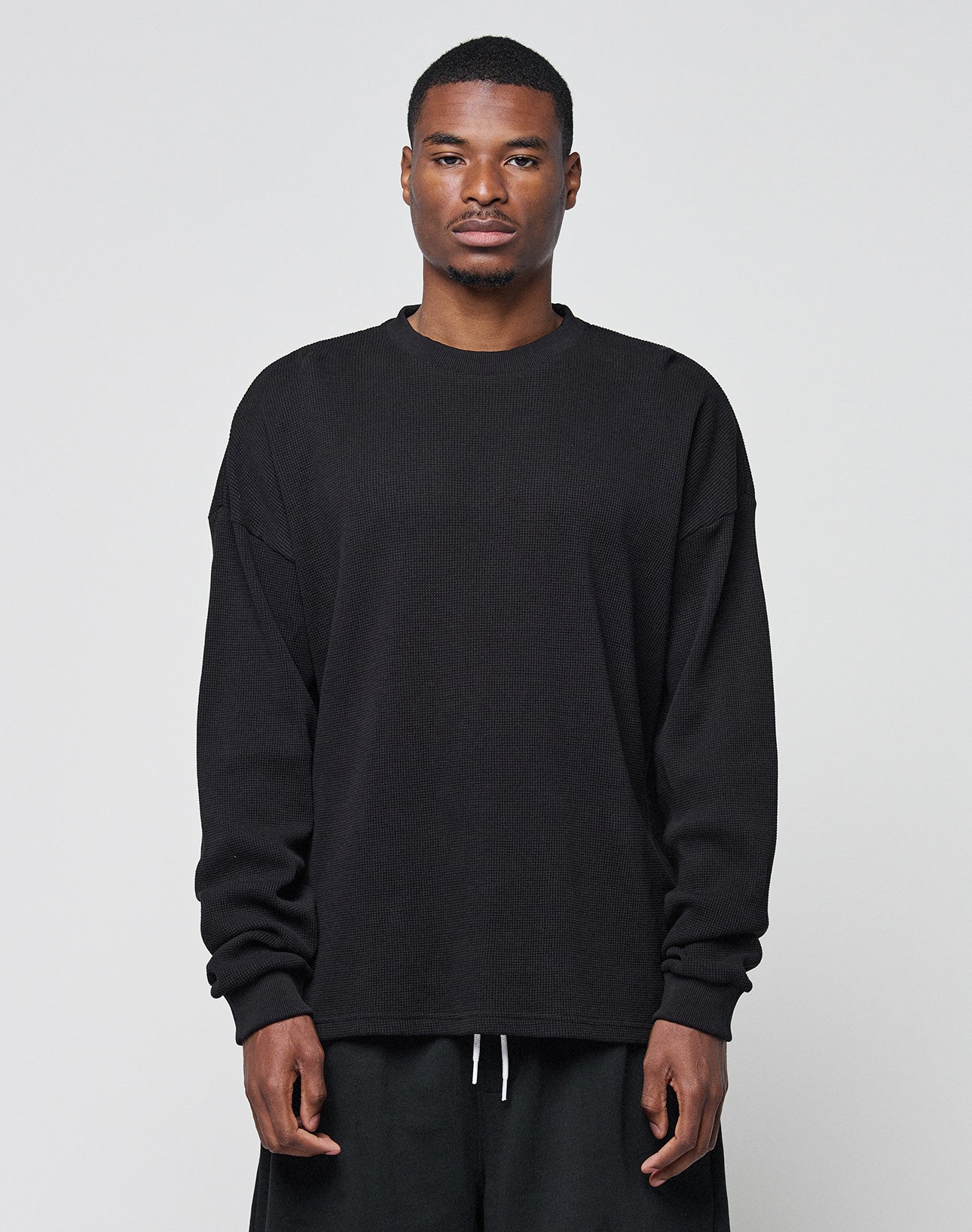 A man faces forward against a plain light gray background, wearing the LIVE FAST DIE YOUNG Blank Waffle Longsleeve in black, made from 100% Baumwolle, styled with dark pants and a neutral expression.