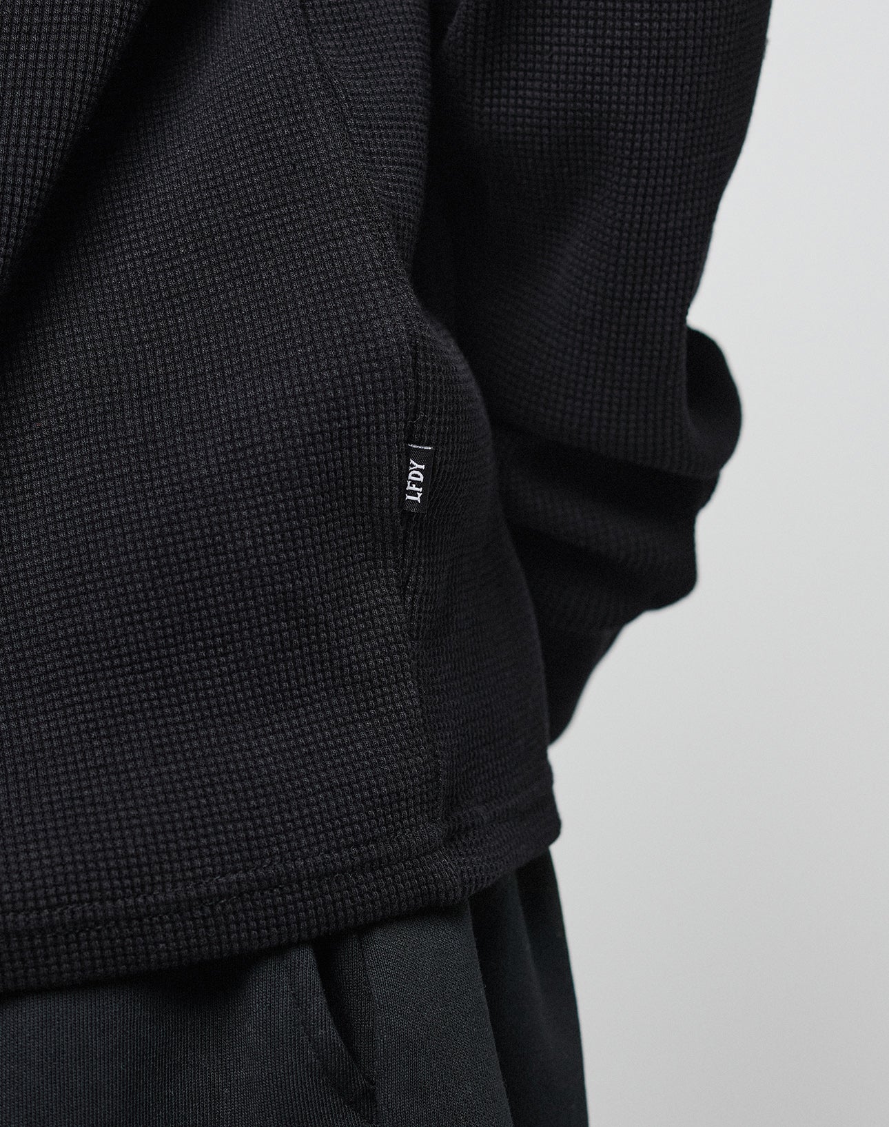 Close-up of a person in the Blank Waffle Longsleeve by LIVE FAST DIE YOUNG, an oversize-fit, textured black sweater made from 100% Baumwolle, paired with black pants. Adpt tag is sewn into the side seam against a light background.