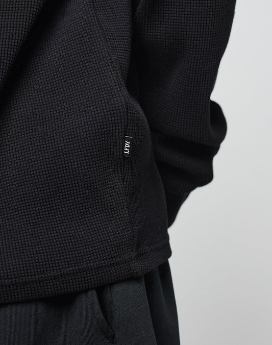 Close-up of a person in the Blank Waffle Longsleeve by LIVE FAST DIE YOUNG, an oversize-fit, textured black sweater made from 100% Baumwolle, paired with black pants. Adpt tag is sewn into the side seam against a light background.