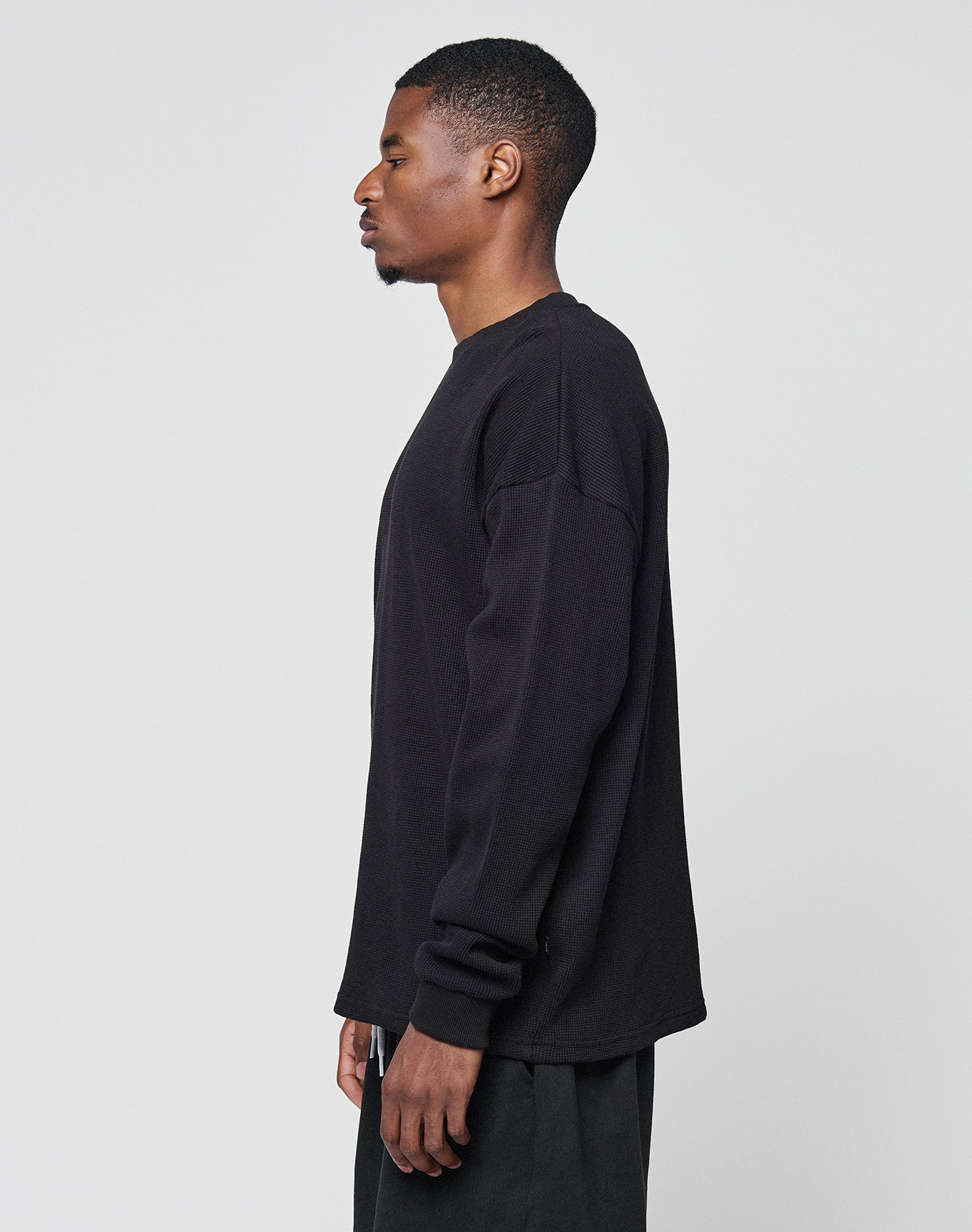 A man with short hair stands in profile against a plain, light background, wearing the LIVE FAST DIE YOUNG Blank Waffle Longsleeve in an Oversize Fit and dark pants.