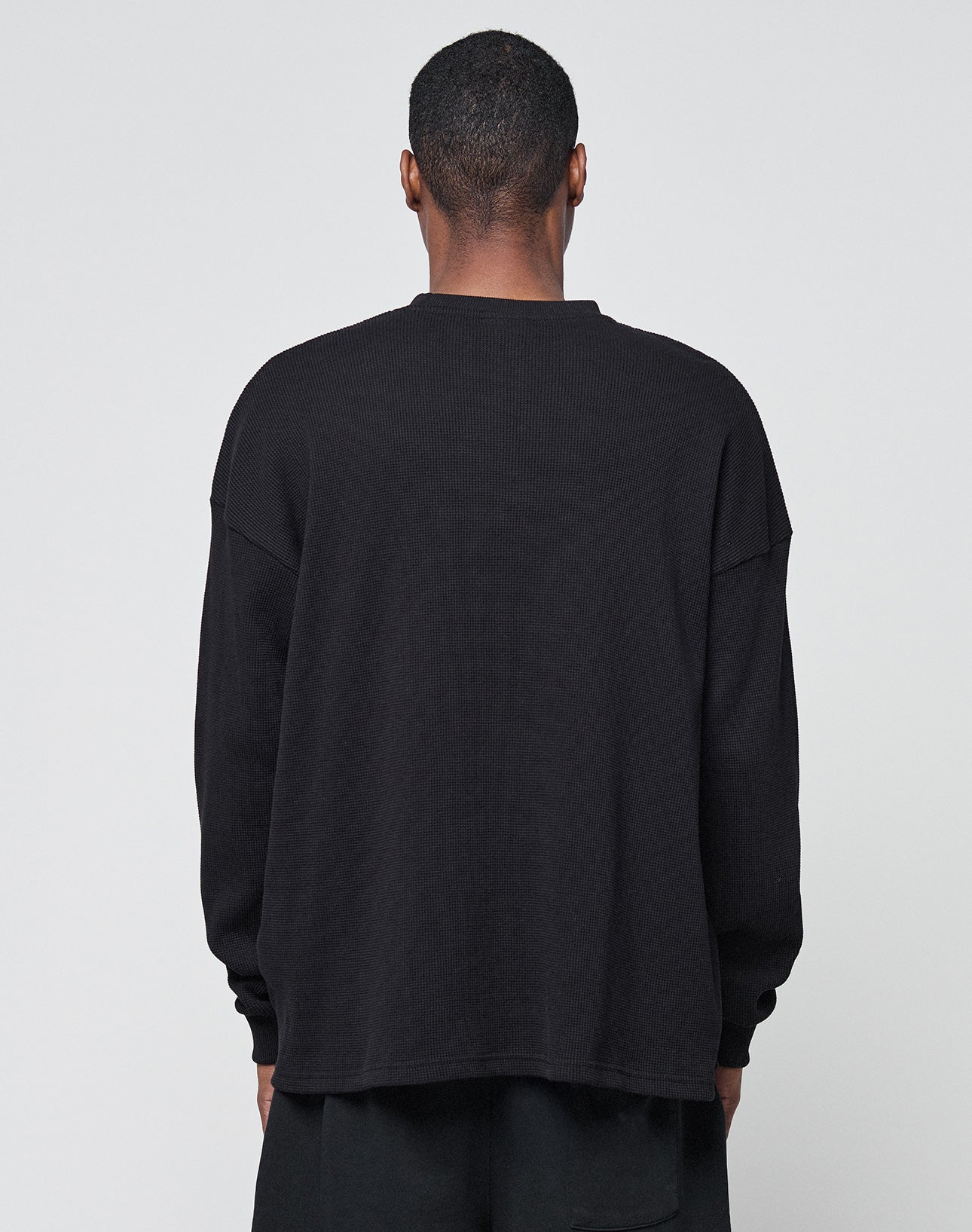 A person with short hair faces away from the camera, wearing LIVE FAST DIE YOUNGs Blank Waffle Longsleeve—an oversize black 100% Baumwolle shirt—and black pants, set against a plain light gray background.