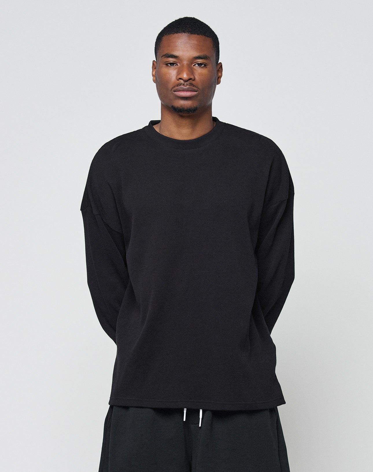 A man stands against a plain light gray background, wearing the LIVE FAST DIE YOUNG Blank Waffle Longsleeve in black and matching black pants. He faces the camera with a neutral expression, arms relaxed at his sides.
