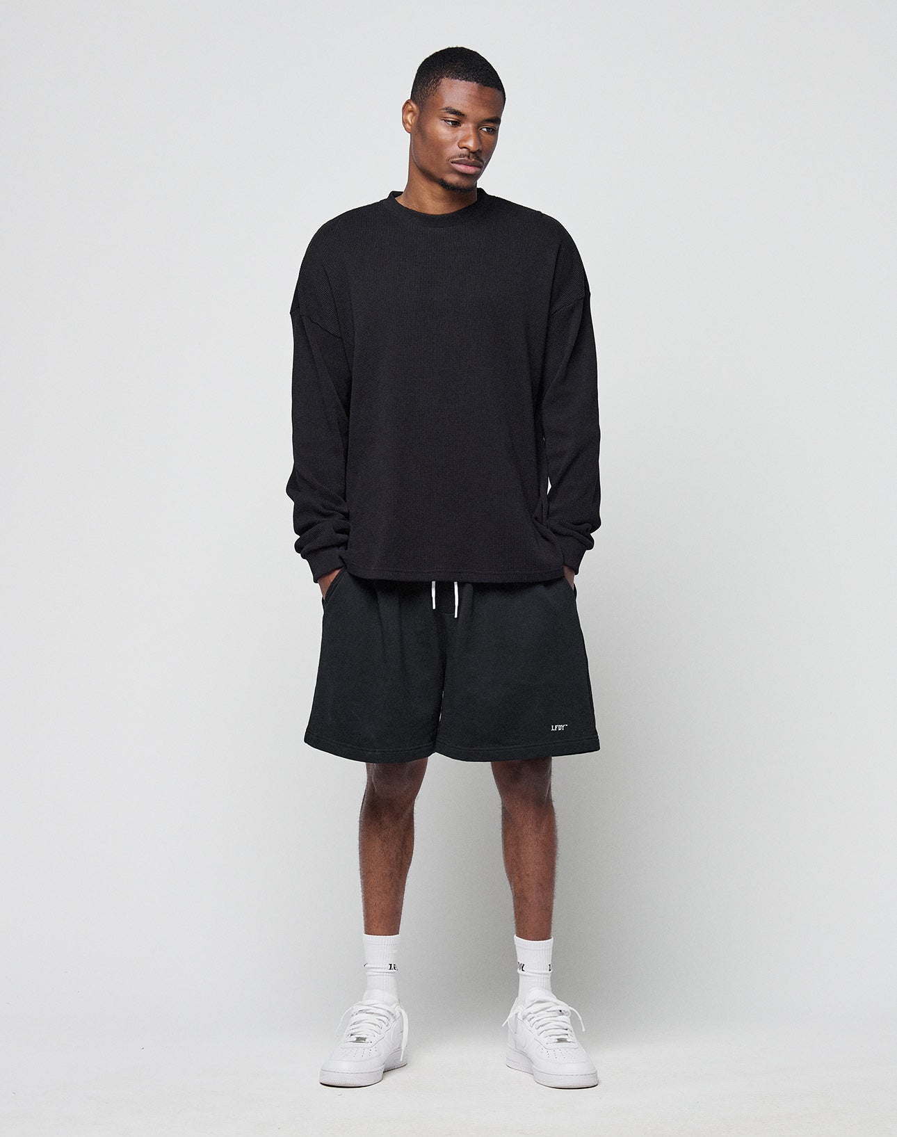 A man stands against a white background, wearing the LIVE FAST DIE YOUNG Blank Waffle Longsleeve in black (100% Baumwolle), black shorts with white drawstrings, white socks, and sneakers. He looks slightly downward with his hands at his sides.