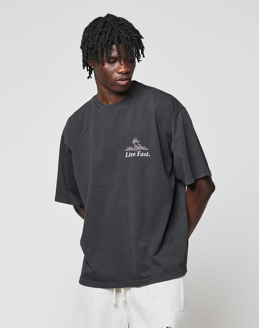 A person with short, twisted hair wears the LIVE FAST DIE YOUNG LFDY Praying Tee in an oversize fit with front print, paired with light gray pants, standing against a plain light background.