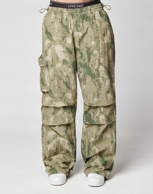The LIVE FAST DIE YOUNG Live Fast Nylon Pants feature a loose oversize fit, elastic cord ankles, green/beige camo print, and a waistband with LIVE FAST print. Shown with white sneakers on a plain white background.