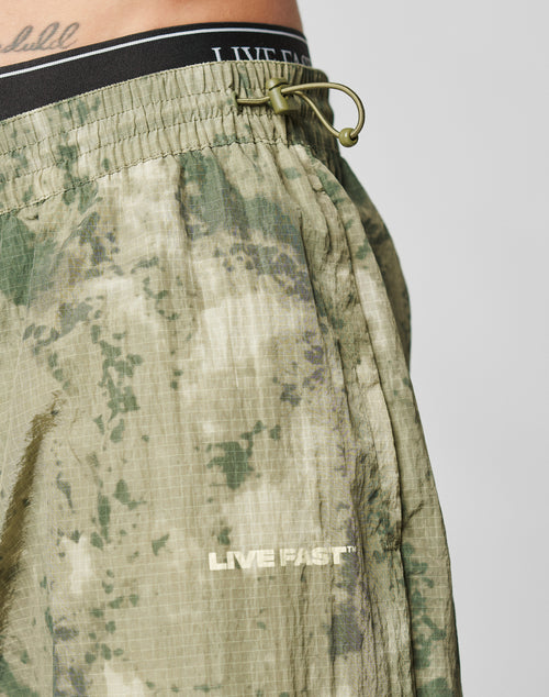 Close-up of a person wearing LIVE FAST DIE YOUNG’s Live Fast Nylon Pants featuring green tie-dye, LIVE FAST printed in white by the pocket, an elastic waistband with black cord, and an oversized fit for comfort.