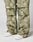 A close-up of a person wearing LIVE FAST DIE YOUNGs Live Fast Nylon Pants in green and beige camouflage with wide legs, cargotasche, layered panels, and an oversize fit, paired with white sneakers against a plain light background.