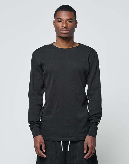 A man stands facing forward against a plain light background, wearing the LIVE FAST DIE YOUNG Basic Regular Rib Longsleeve in black with matching black shorts, his expression neutral and arms relaxed at his sides.