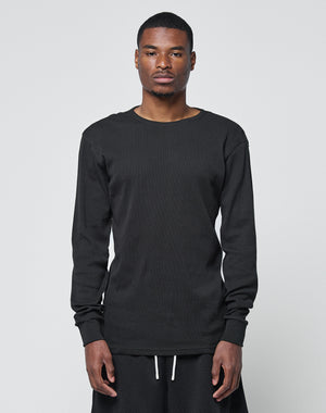 A man stands facing forward against a plain light background, wearing the LIVE FAST DIE YOUNG Basic Regular Rib Longsleeve in black with matching black shorts, his expression neutral and arms relaxed at his sides.