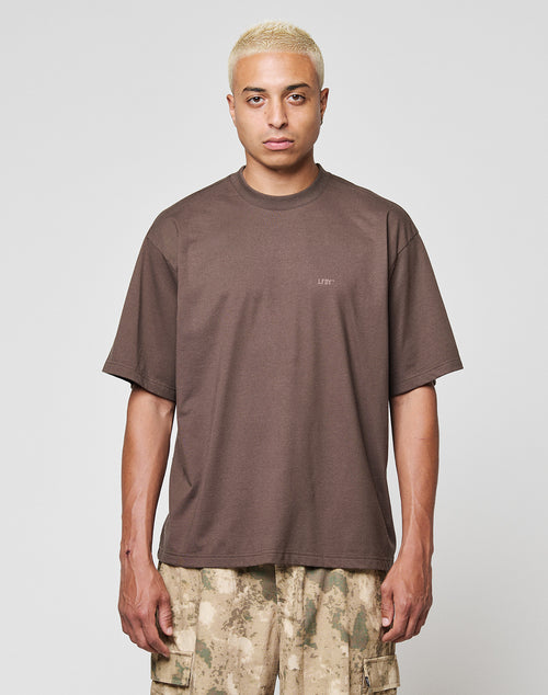 A person with short bleached blond hair wears a loose brown LFDY Flash Tee by LIVE FAST DIE YOUNG and beige camouflage cargo pants, standing against a plain light gray background, facing forward with a neutral expression.