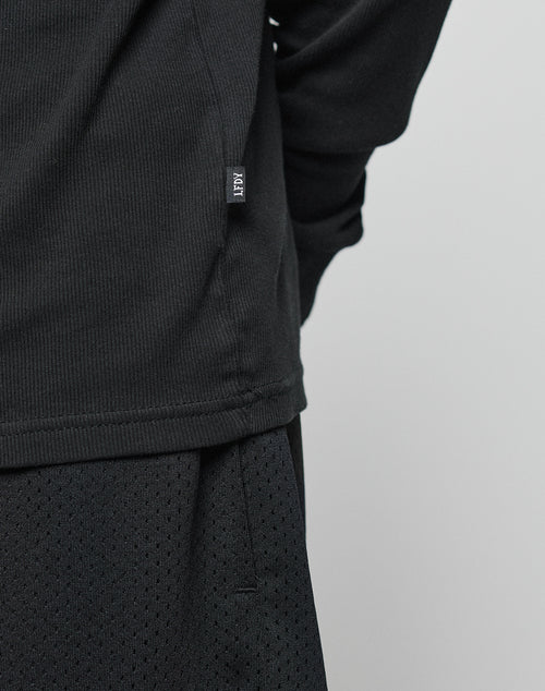 A close-up of a person wearing the LIVE FAST DIE YOUNG Basic Regular Rib Longsleeve in black with a side-seam tag, paired with black mesh athletic shorts against a plain light background.