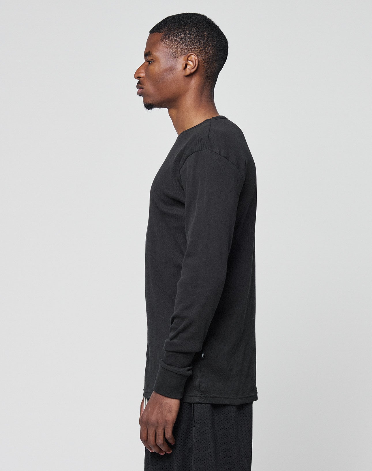 A man with short hair stands in profile, facing left, wearing the LIVE FAST DIE YOUNG Basic Regular Rib Longsleeve and black shorts, both regular fit, against a plain light gray background.