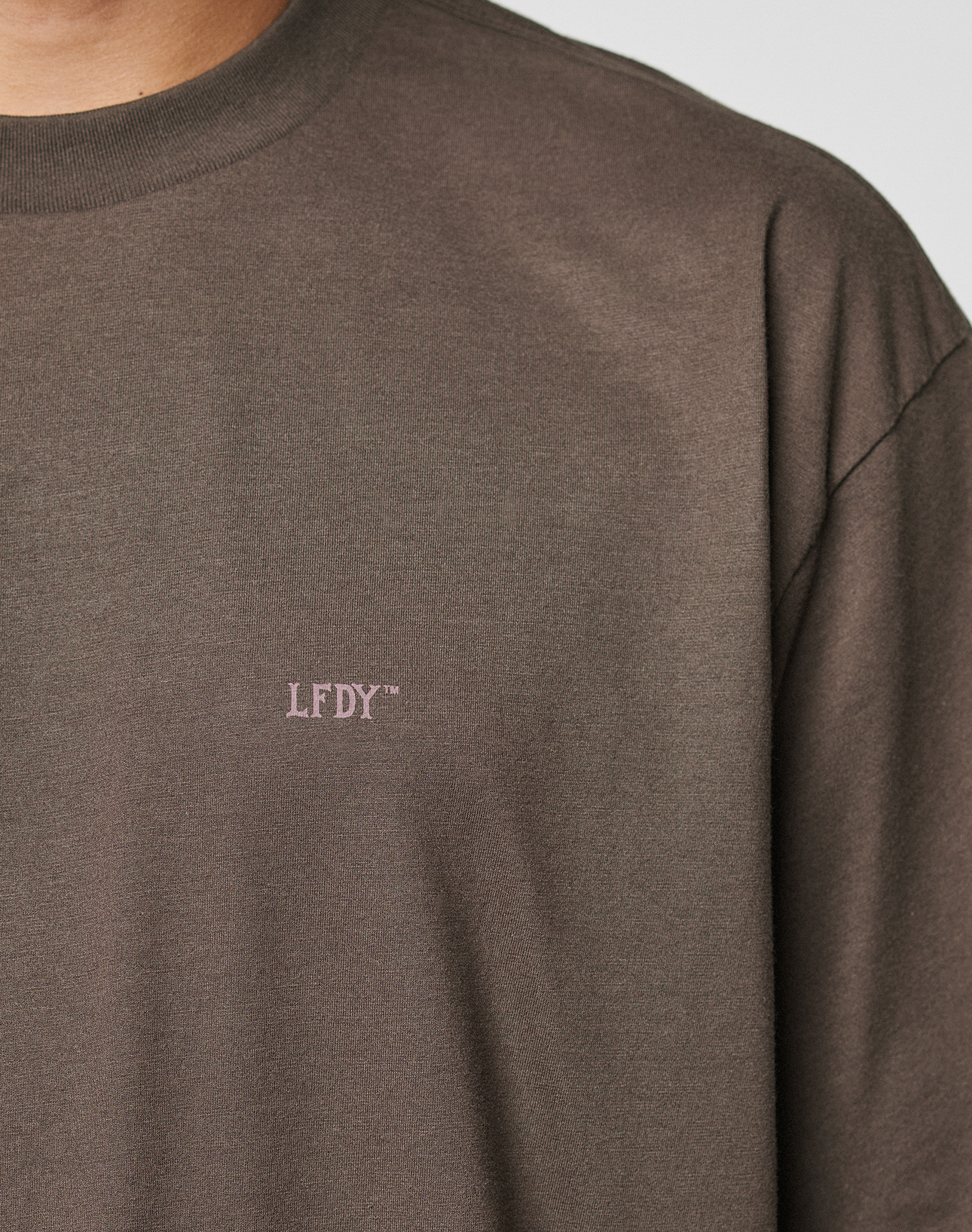 A close-up shows part of the neck, shoulder, and upper chest of a person wearing the LIVE FAST DIE YOUNG LFDY Flash Tee, a plain brown oversize fit t-shirt with small light-colored "LFDY" text on the chest.