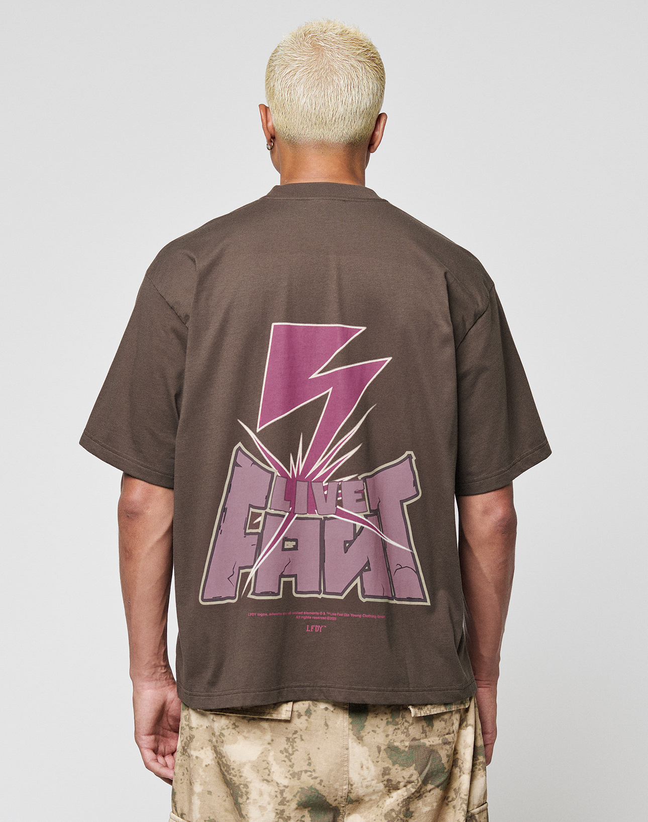 A person with short bleach-blond hair wears the LFDY Flash Tee by LIVE FAST DIE YOUNG, an oversize brown cotton-slub t-shirt with a bold purple lightning bolt and "LIVE FAST" print on front and back, paired with brown-beige camo shorts. | model