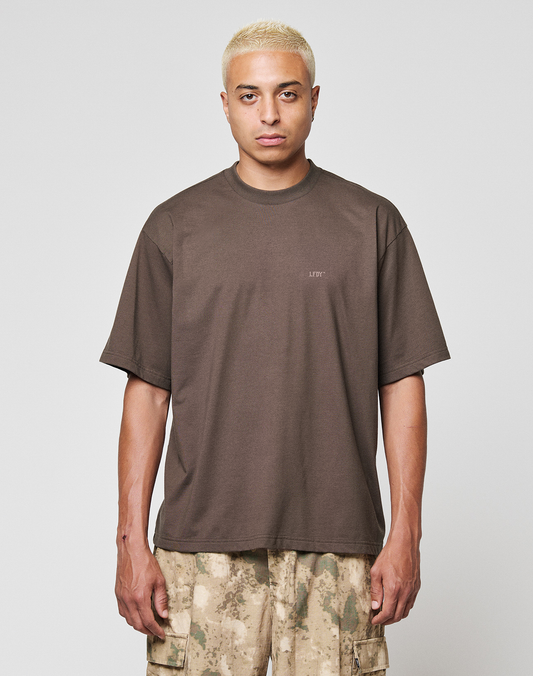 A person with short bleached blonde hair wears the LIVE FAST DIE YOUNG LFDY Flash Tee, a loose-fitting dark brown Oversize Fit top made from Cotton-Slub Stoff, and beige camouflage cargo shorts, standing against a light gray background.