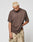 A person with short bleached blond hair wears a loose LFDY Flash Tee by LIVE FAST DIE YOUNG and tan camouflage cargo pants, standing against a plain light background and looking down while gripping their shirt.