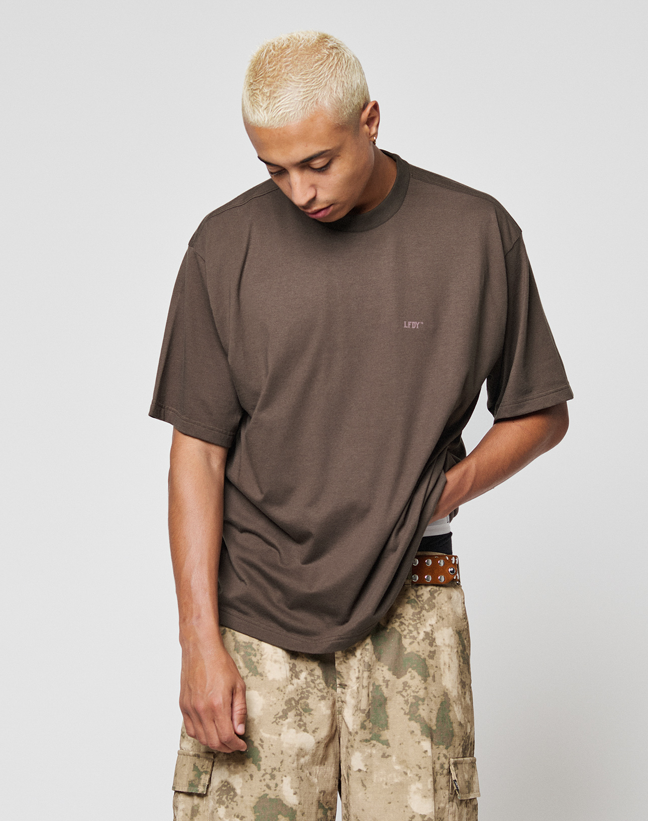 A person with short, bleached blond hair looks down while wearing the LIVE FAST DIE YOUNG LFDY Flash Tee in brown and beige camouflage cargo pants, set against a plain white background.