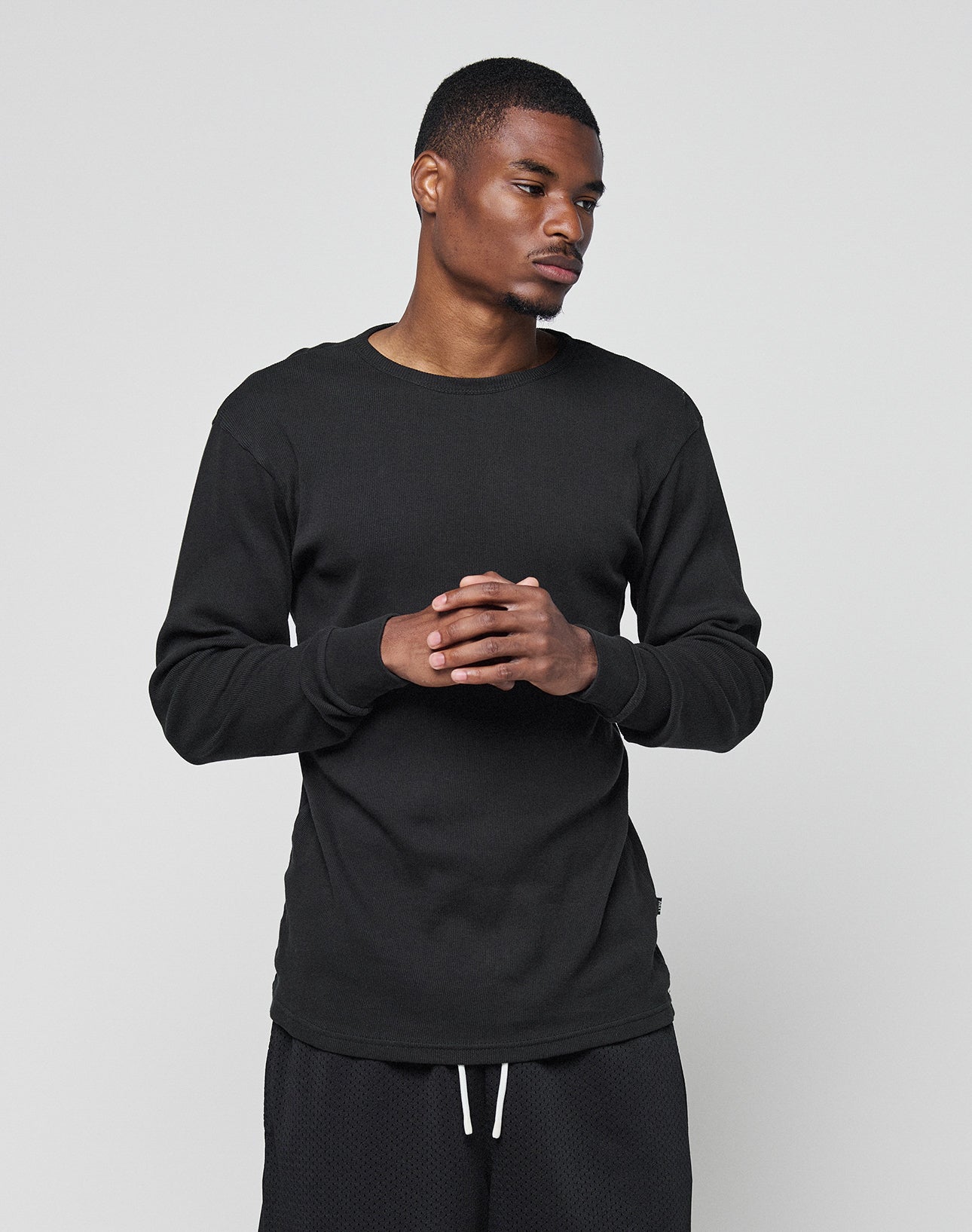 A man wearing the LIVE FAST DIE YOUNG Basic Regular Rib Longsleeve and black shorts in a regular fit stands against a light gray background with his hands clasped, looking slightly to the side.