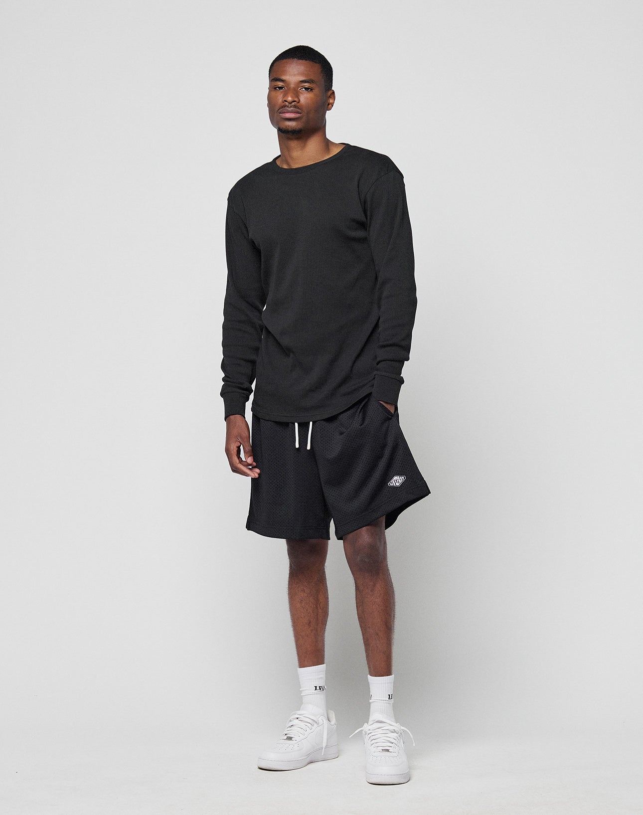 A man stands against a light background, confidently looking at the camera with one hand in his pocket. He wears the LIVE FAST DIE YOUNG Basic Regular Rib Longsleeve in black, paired with black shorts, white crew socks, and white sneakers.