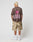 A person with short blonde hair faces away, wearing the oversized LIVE FAST DIE YOUNG LFDY Flash Tee featuring a pink lightning bolt and LIFE FAST back print, camouflage cargo shorts, white socks, and white sneakers.