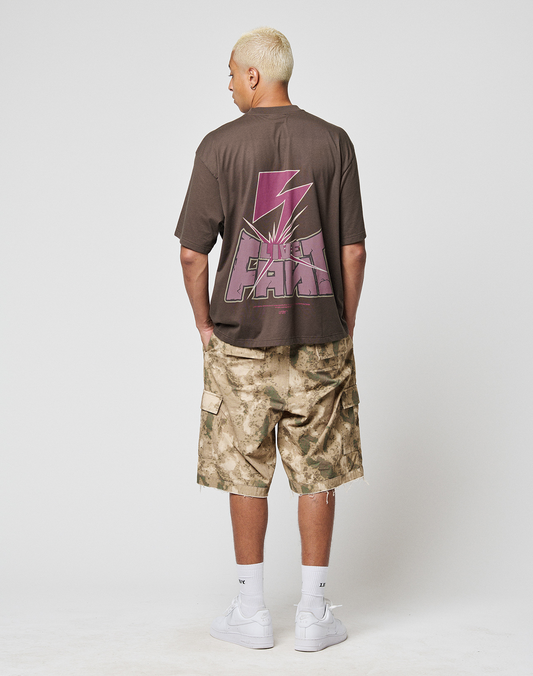 A person with short blond hair faces away, wearing the LIVE FAST DIE YOUNG LFDY Flash Tee—an oversized dark cotton-slub shirt with bold pink graphics and “UNDER PEACE” prints—styled with camo shorts, white socks, and sneakers against a plain background.
