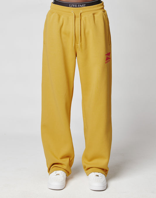 A person wears the LIVE FAST DIE YOUNG Live Fast Sweatsuit Jogger in mustard yellow with an oversize fit, elastic waistband, red logo on the left thigh, white sneakers, and a black LIVE FAST waistband against a plain white background.