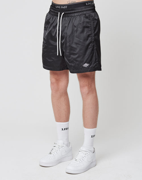 A person wearing LIVE FAST DIE YOUNGs Basic Satin Shorts in black with a white waistband and drawstring, paired with white crew socks and sneakers, stands against a plain white background.
