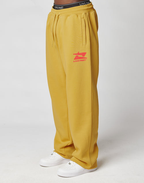 A person wears the LIVE FAST DIE YOUNG Live Fast Sweatsuit Jogger in mustard yellow with a red LIVE FAST logo on the left thigh, Elastik-Bund, oversize fit, white sneakers, and a visible black waistband. Background is plain white.