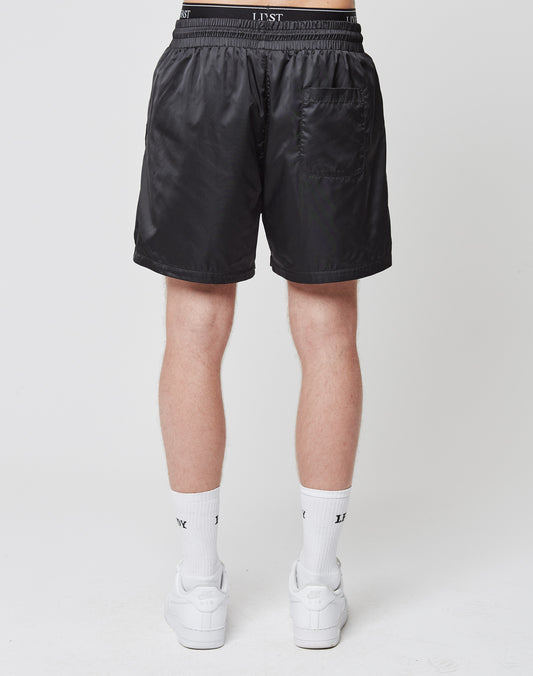A person wearing LIVE FAST DIE YOUNGs Basic Satin Shorts, featuring an elastic waistband, LST logo patch, and back pocket, stands with their back to the camera. The shorts are paired with white socks and sneakers on a plain white background.