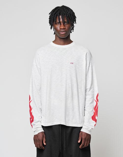 A young man with short dreadlocks models the LIVE FAST DIE YOUNG LFDY We Walked Longsleeve—an oversize white shirt with red flames on the sleeves and small red chest text, made from 100% Baumwolle, against a plain light gray background.