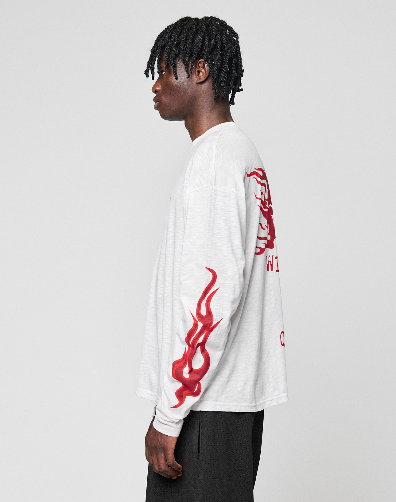 A person in an oversize LFDY We Walked Longsleeve by LIVE FAST DIE YOUNG, with red flame graphics and a bold logo frontprint, stands in profile. The shirt is made from soft cotton. Their hair is styled in short twists.