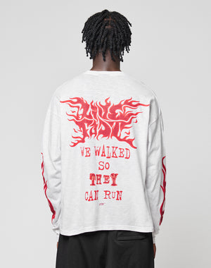 A person with braided hair, seen from behind, wears the LFDY We Walked Longsleeve by LIVE FAST DIE YOUNG—an oversized white 100% Baumwolle shirt with bold red “WE WALKED SO THEY CAN RUN.” text and flame designs on the back and sleeves.