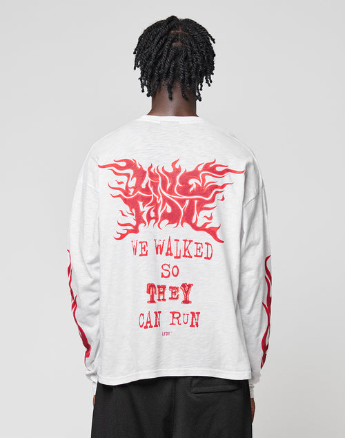 A person with braided hair, seen from behind, wears the LFDY We Walked Longsleeve by LIVE FAST DIE YOUNG—an oversized white 100% Baumwolle shirt with bold red “WE WALKED SO THEY CAN RUN.” text and flame designs on the back and sleeves.
