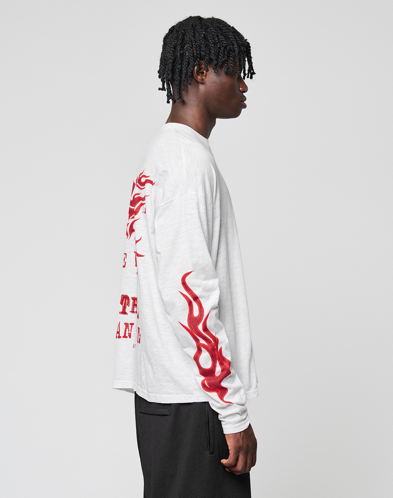 A person with short twisted hair stands sideways, wearing the LIVE FAST DIE YOUNG LFDY We Walked Longsleeve—an oversized white cotton shirt featuring red flame sleeve designs and a bold back logo print—paired with black pants.