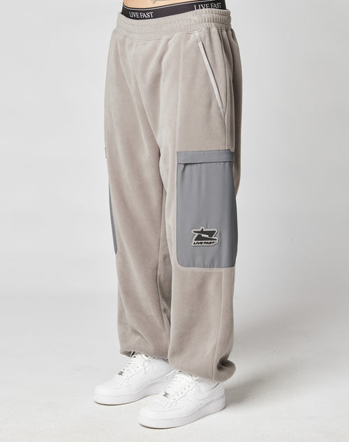 A person wearing LIVE FAST DIE YOUNGs Live Fast Fleece Pants—loose-fitting, light gray with wide fit, gray nylon pockets, and elastic waistband—paired with white Nike sneakers, stands against a plain background.
