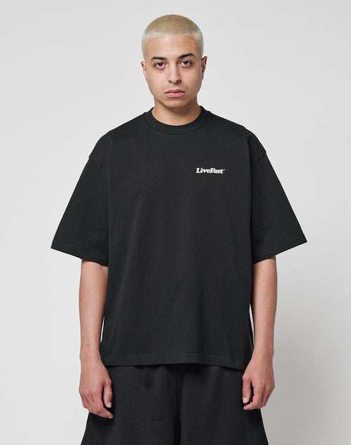 A person with short, bleached blonde hair wears the LIVE FAST DIE YOUNG Basic 280 Tee in black, featuring a small white LiveFast chest print and an oversize fit, paired with black shorts against a plain white background.