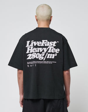 A person with short bleached hair wears a black LIVE FAST DIE YOUNG Basic 280 Tee aus Baumwolle, showcasing bold white “Live Fast Heavy Tee 280g/m²” text with Front- und Backprint icons. The background is plain and light-colored.