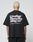 A person with short bleached hair wears a black LIVE FAST DIE YOUNG Basic 280 Tee aus Baumwolle, showcasing bold white “Live Fast Heavy Tee 280g/m²” text with Front- und Backprint icons. The background is plain and light-colored.