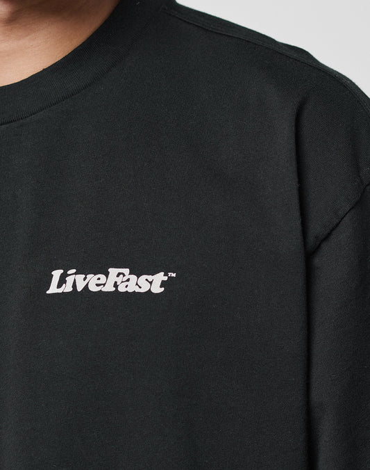 Close-up of a person wearing the LIVE FAST DIE YOUNG Basic 280 Tee in black, featuring white LiveFast™ text on the left chest. Only part of the face and upper torso are visible.