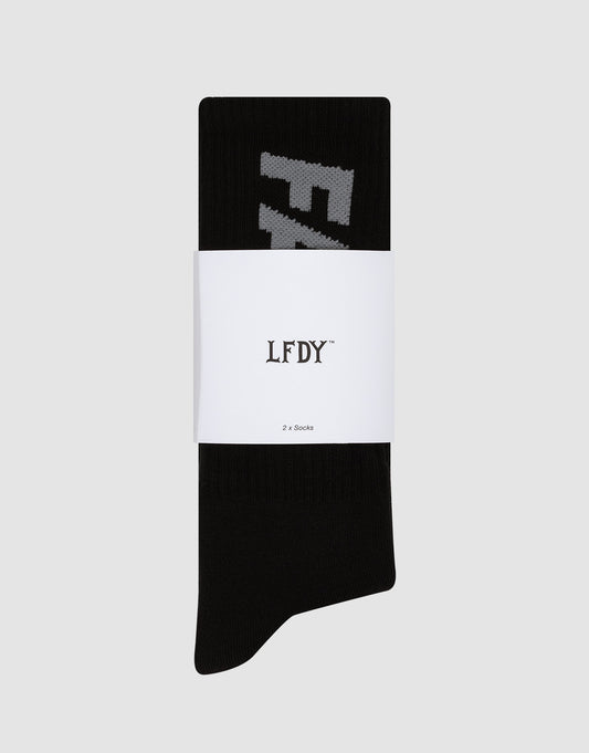 A pair of black LIVE FAST DIE YOUNG Basic Typo Socks (2 Pack), folded with a white LFDY Logo and 2er Set paper band, rests on a light gray background. Partial white lettering appears on the socks.