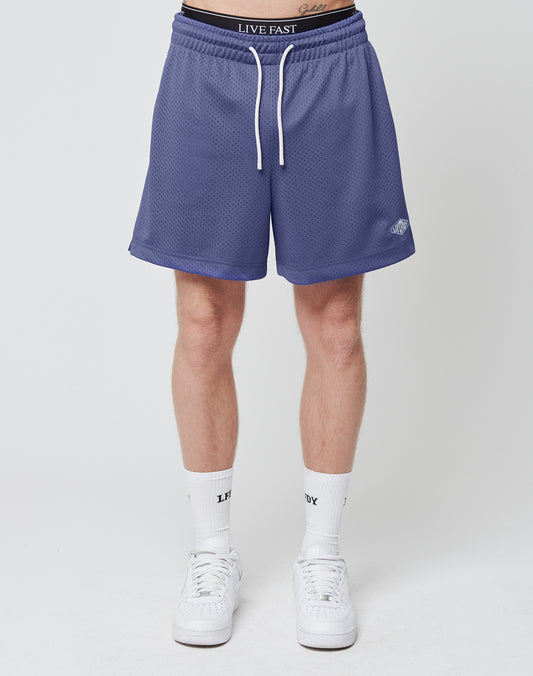 A person wears LIVE FAST DIE YOUNG Basic Mesh Shorts with an elastic waistband and subtle logo patch, paired with white high socks and sneakers, standing against a plain white background. Only their lower torso and legs are shown.